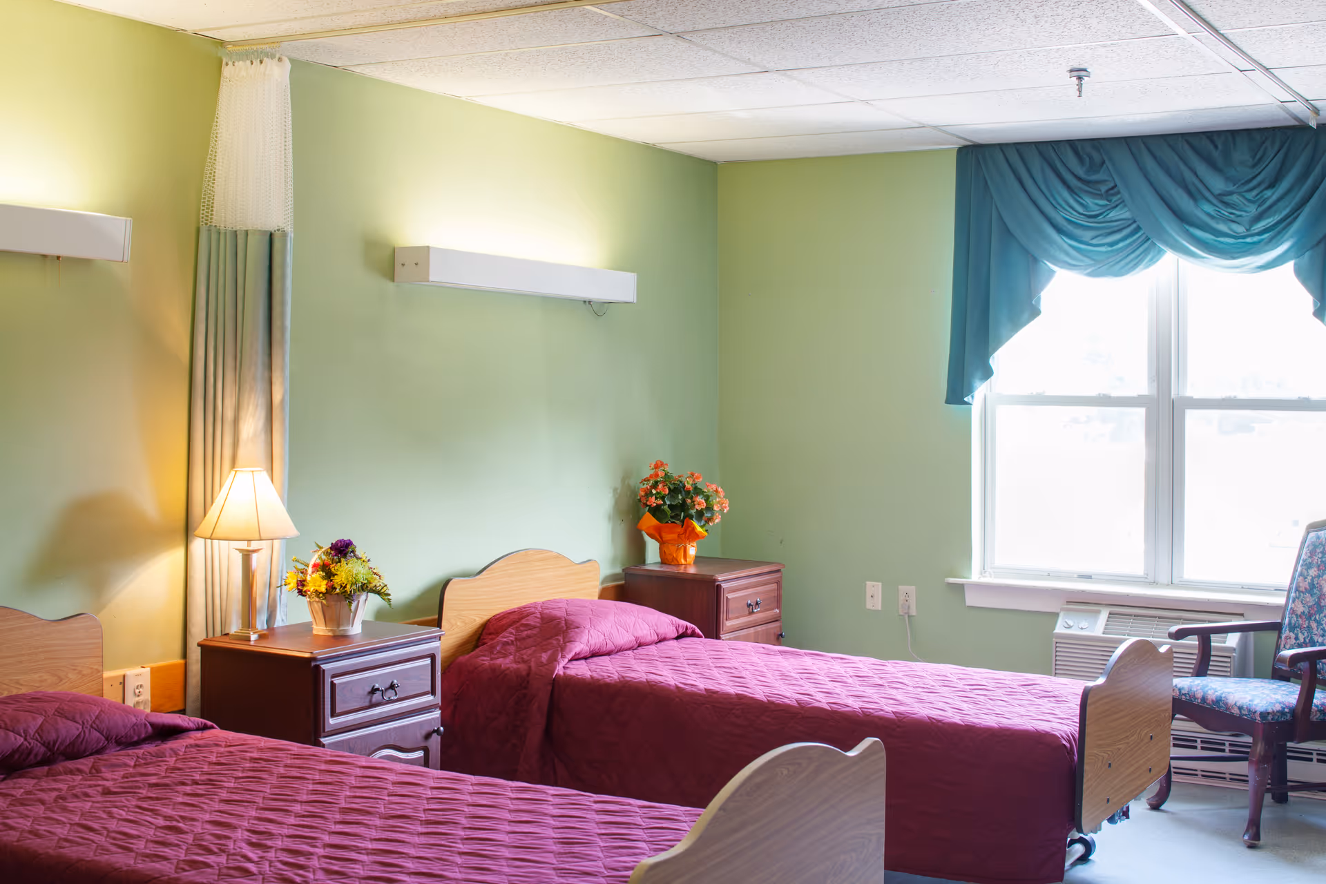 A bright room with two single beds covered in matching maroon quilts. Between the beds is a wooden nightstand with a lamp and a small flower arrangement. Another nightstand with a flower pot is against the far wall. The walls are painted light green, and a window with teal drapes lets in natural light. A floral upholstered chair is positioned near the window.