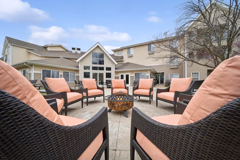 Outdoor seating area at College View Manor featuring a circular arrangement of cushioned wicker chairs around a central fire pit, with a multi-story beige building and leafless tree in the background under a partly cloudy sky.