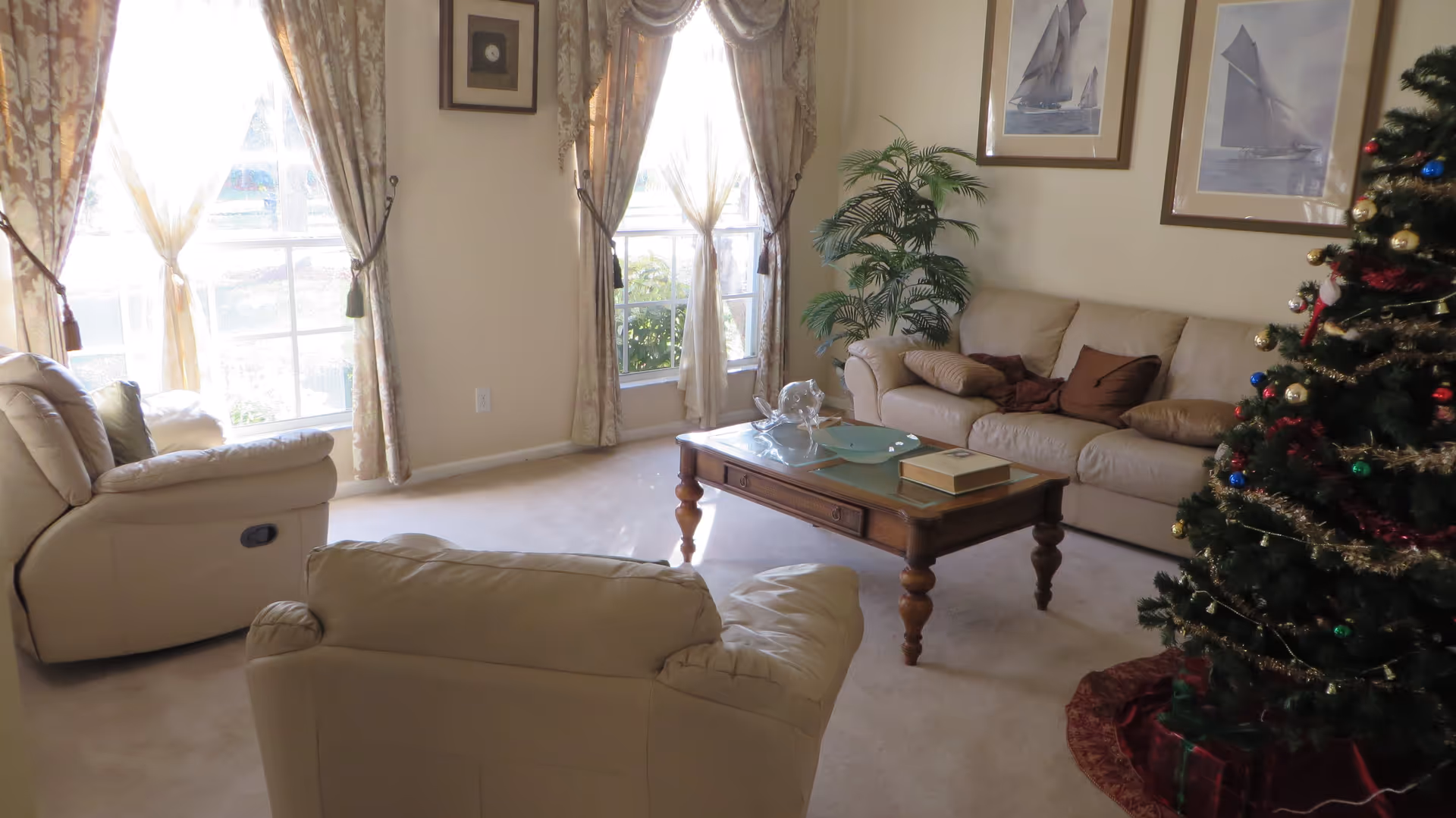 A cozy living room with beige leather sofas and armchairs, a wooden coffee table with decorative items, large windows with patterned curtains letting in natural light, framed pictures of sailboats on the wall, a potted plant in the corner, and a decorated Christmas tree with wrapped presents underneath.