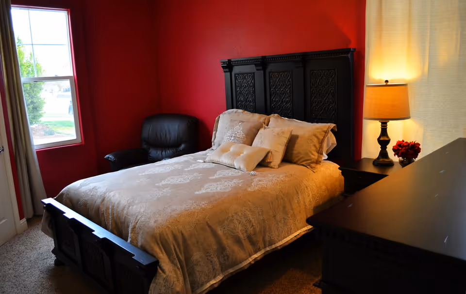 A cozy bedroom with red walls featuring a large bed with beige patterned bedding and multiple pillows. There is a black leather armchair near a window with beige curtains, a black nightstand with a lamp and a small vase of red flowers, and a black dresser in the foreground.