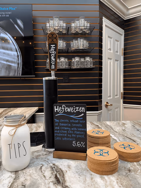 A countertop with a beer tap labeled Hefeweizen, a small chalkboard sign describing the beer, a jar labeled TIPS, and stacks of round coasters with a blue flower design. Behind the counter are shelves holding glass mugs and a closed white door.