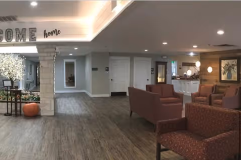 A spacious and well-lit common area in a senior living facility with wood-style flooring, several upholstered armchairs arranged for conversation, a stone pillar, and a decorative sign on the ceiling that reads 'WELCOME home'. There is a small indoor garden area with a pumpkin and a white flowering plant, and a reception desk with a lamp and framed artwork on the wall behind it.