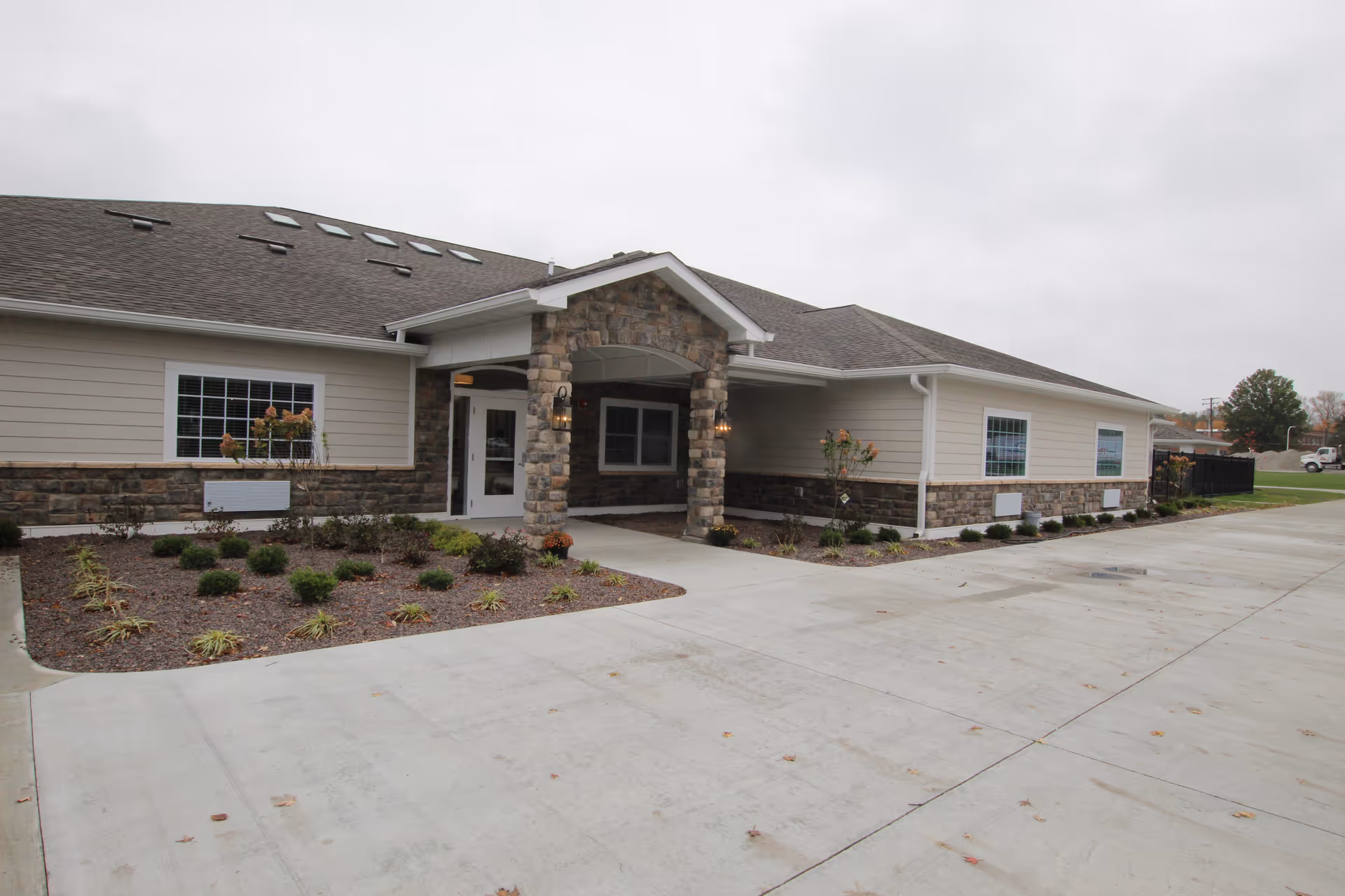 Exterior view of a single-story building with beige siding and stone accents, featuring a covered entrance with stone pillars and a concrete driveway in front. The sky is overcast and there are small landscaped areas with shrubs and plants near the building.