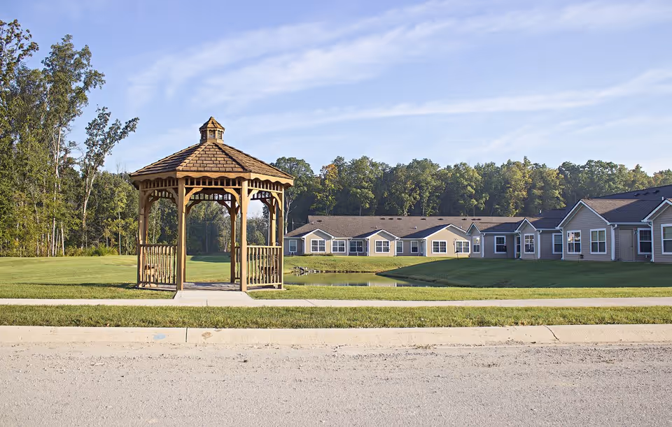 A wooden gazebo on a grassy lawn with a paved sidewalk in front. Behind the gazebo, there is a small pond and a row of single-story residential buildings with beige siding and white trim. Trees and a blue sky with light clouds are visible in the background.