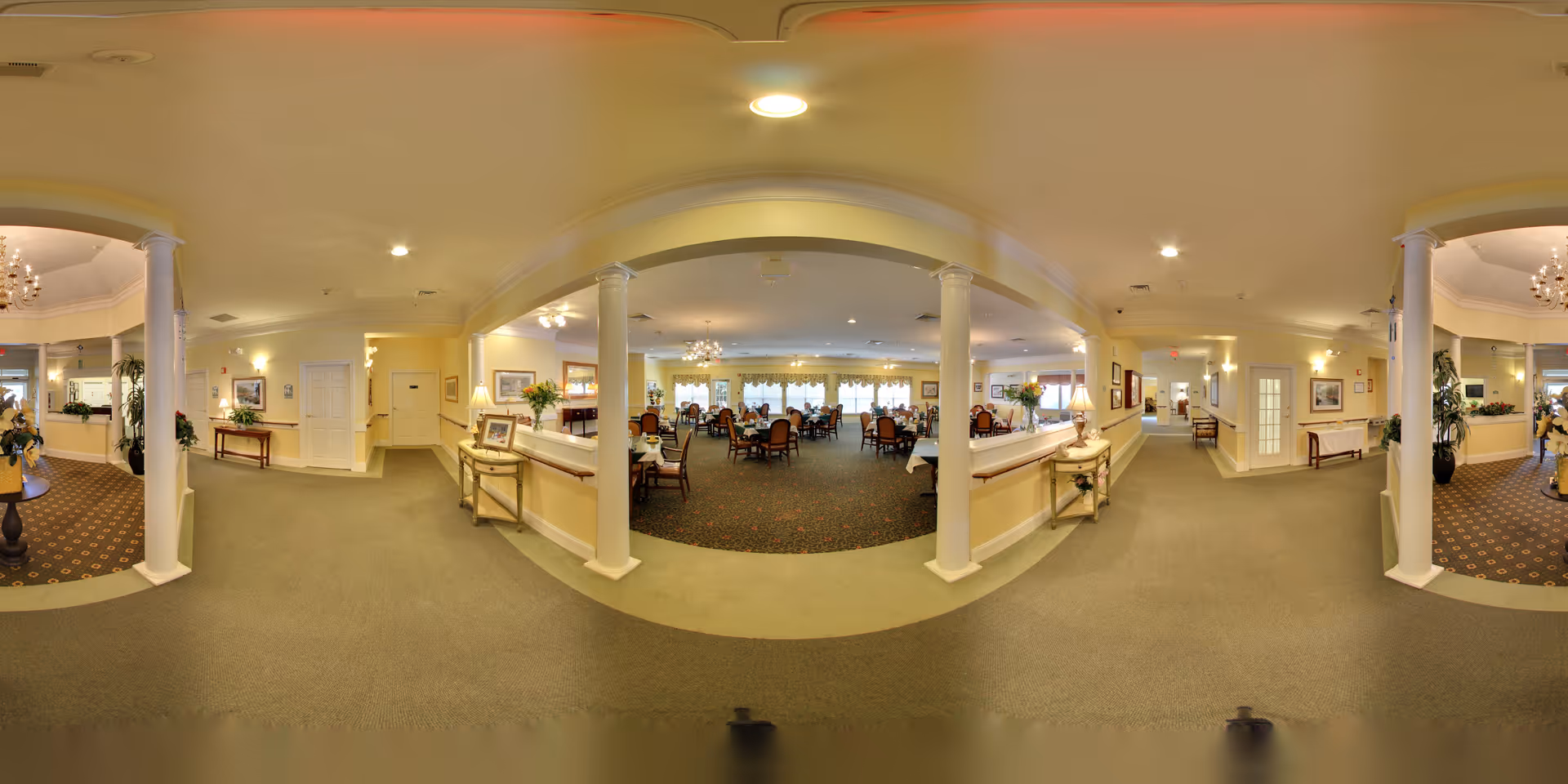 Panoramic view of a senior living facility's interior showing a spacious dining area with multiple tables and chairs, elegant chandeliers, columns, and a carpeted floor. The area is well-lit with warm lighting and decorated with plants and framed pictures on the walls.