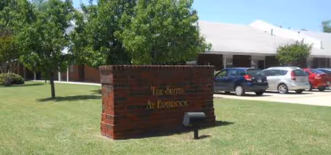 Front exterior of The Suites at Elmbrook with brick sign and parked cars.