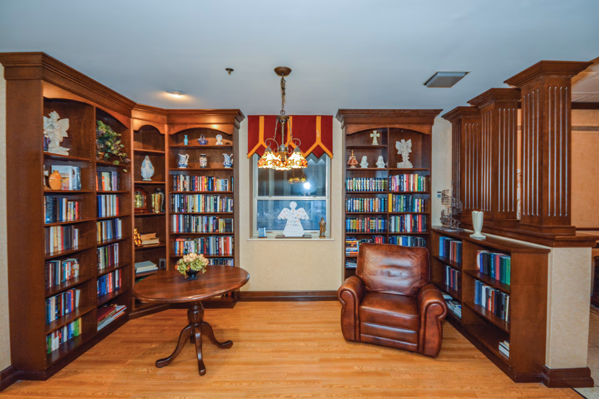 Cozy library-style common room with wooden bookshelves, a round table, a leather armchair, and decorative accents.