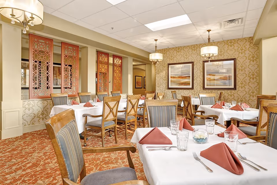 A well-lit dining room with several tables covered in white tablecloths, each set with folded red napkins, glasses, and silverware. The room features patterned carpet, decorative wallpaper, framed landscape paintings on the wall, and wooden chairs with cushioned seats. There are hanging light fixtures and decorative red panels on one side of the room.