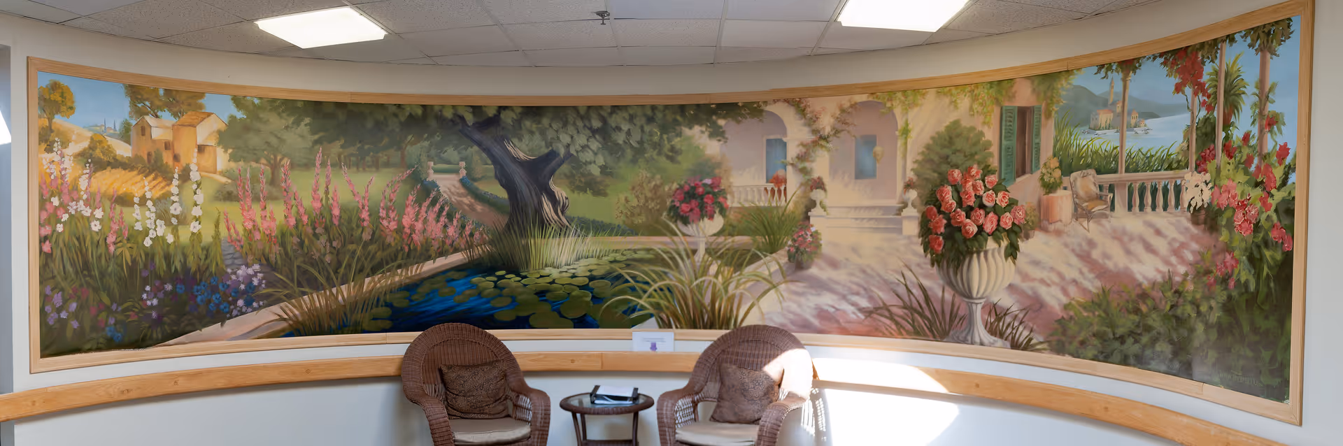 A curved wall mural depicting a serene garden scene with flowers, trees, a pond with lily pads, and a house in the background. In front of the mural are two wicker chairs with cushions and a small round table between them. The setting appears to be a cozy indoor seating area with a wooden bench along the wall.