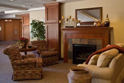 A cozy living room area with a beige sofa adorned with pillows and a red throw blanket, two patterned armchairs, a wooden coffee table with a flower arrangement, a fireplace with a wooden mantle decorated with candles and vases, and a large mirror above the fireplace. There is a potted plant near wooden columns and a hallway in the background.