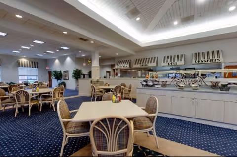 A spacious dining area in a senior living community featuring multiple tables with wicker chairs arranged on a blue carpet with a patterned design. The room has a buffet counter along one wall with sections labeled Deli and Desserts. The ceiling has recessed lighting and a coffered design, and there are plants and windows in the background.