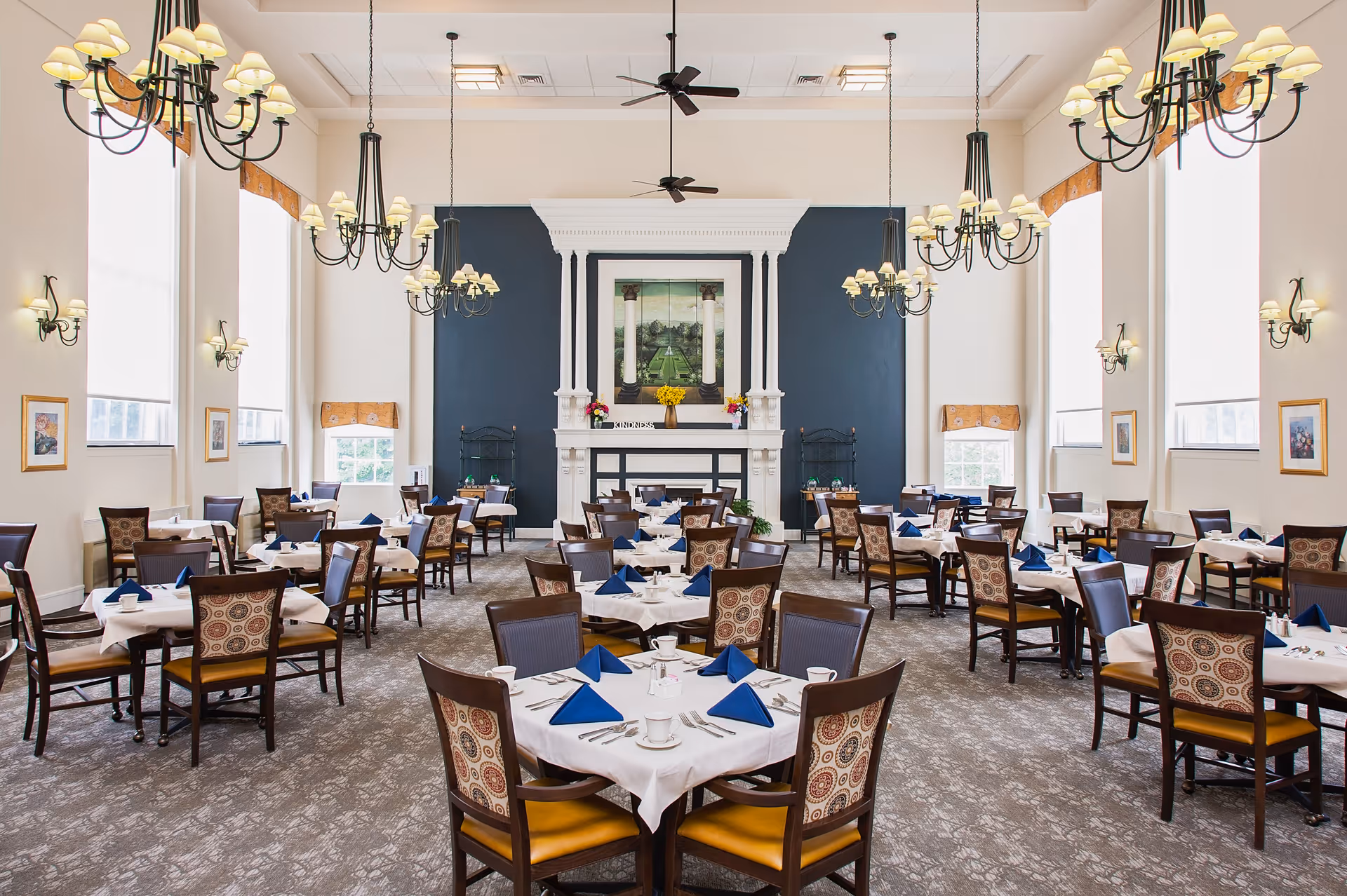 A spacious and elegant dining room with multiple round tables covered in white tablecloths, each set with blue folded napkins, cups, and silverware. The room features high ceilings with several chandeliers and ceiling fans, large windows with light curtains, framed artwork on the walls, and a decorative fireplace with a painting above it at the far end.