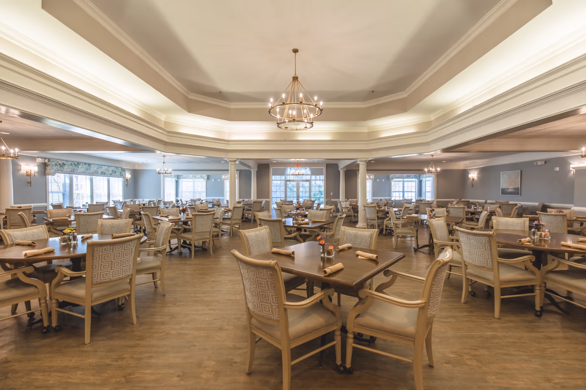 A spacious dining room with multiple wooden tables and cushioned chairs arranged neatly. Each table has rolled napkins and small flower vases. The room features large windows allowing natural light, elegant chandeliers hanging from a tray ceiling, and light-colored walls with decorative molding.