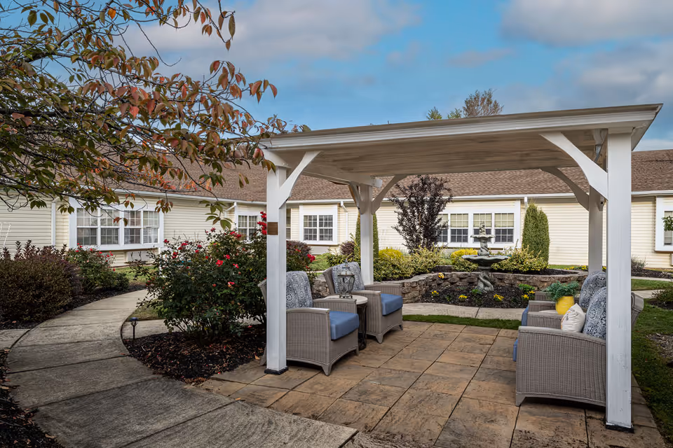 Outdoor seating area at Celebration Villa of York featuring a white pergola with four cushioned wicker chairs arranged around a small table. The area is paved with stone tiles and surrounded by landscaped garden beds with bushes, flowers, and a small fountain. The background shows the exterior of the facility building with beige siding and multiple windows under a partly cloudy sky.
