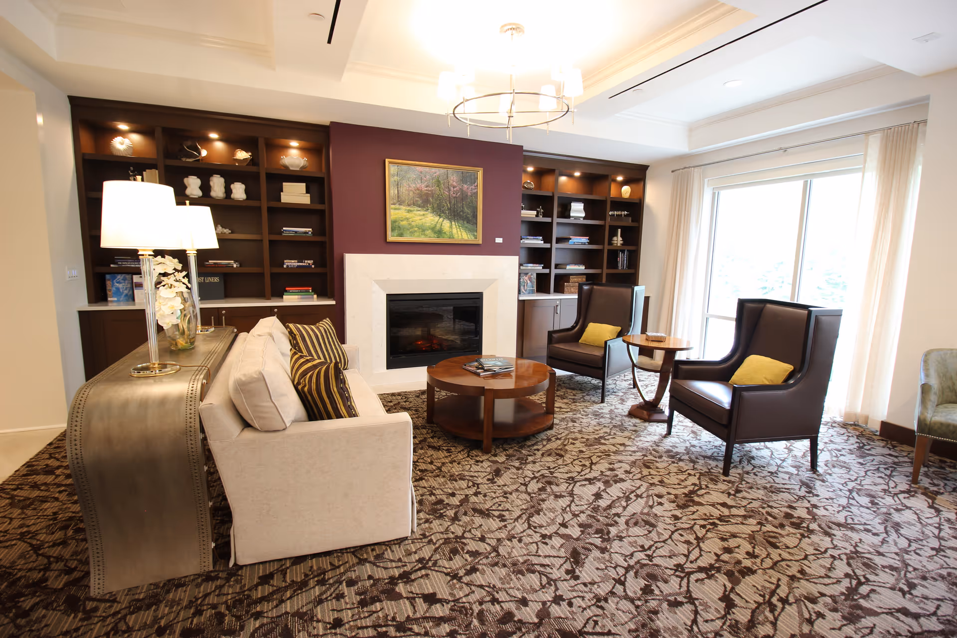 A cozy living room with a beige sofa adorned with striped pillows, two dark brown armchairs with yellow cushions, a round wooden coffee table, and a small side table. The room features a fireplace with a white mantel set against a maroon accent wall, flanked by built-in dark wood shelves filled with decorative items and books. A large window with sheer curtains allows natural light to brighten the space, and a modern chandelier hangs from the ceiling. The floor is covered with a patterned carpet.