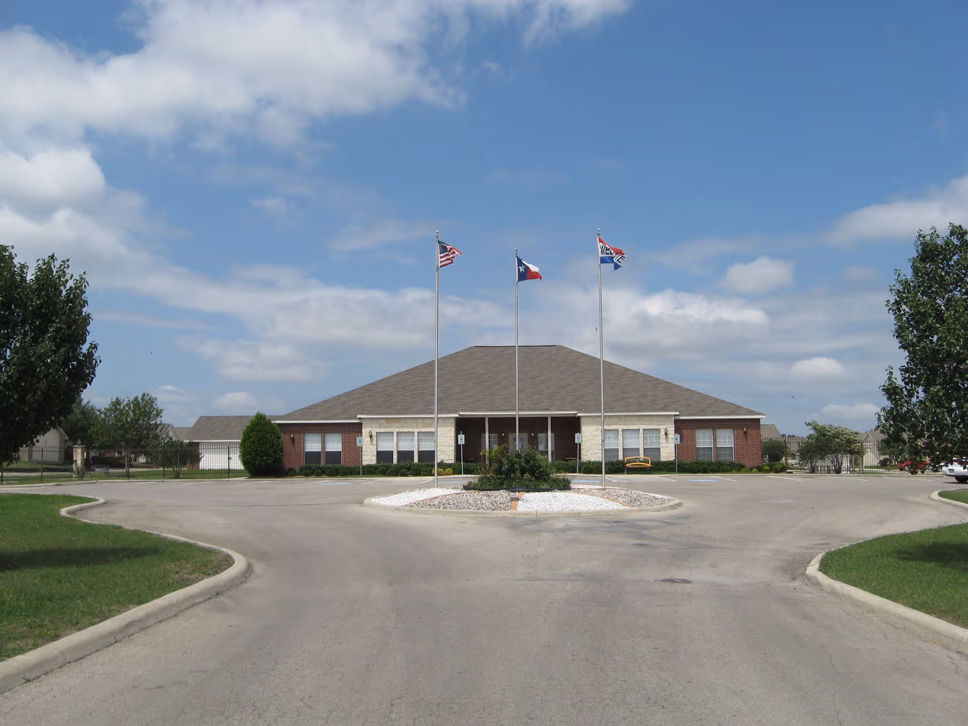 Front exterior view of a single-story brick building with a gray roof, three flagpoles displaying the American flag, Texas state flag, and another flag, surrounded by a circular driveway and green lawns under a partly cloudy sky.