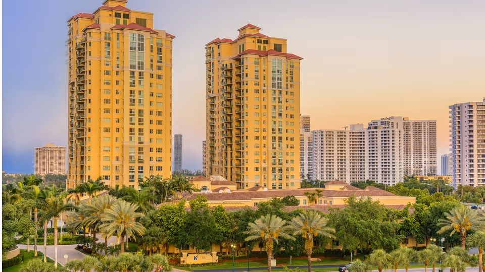 Two tall yellow residential towers surrounded by palm trees with other high-rise buildings in the background at sunset.