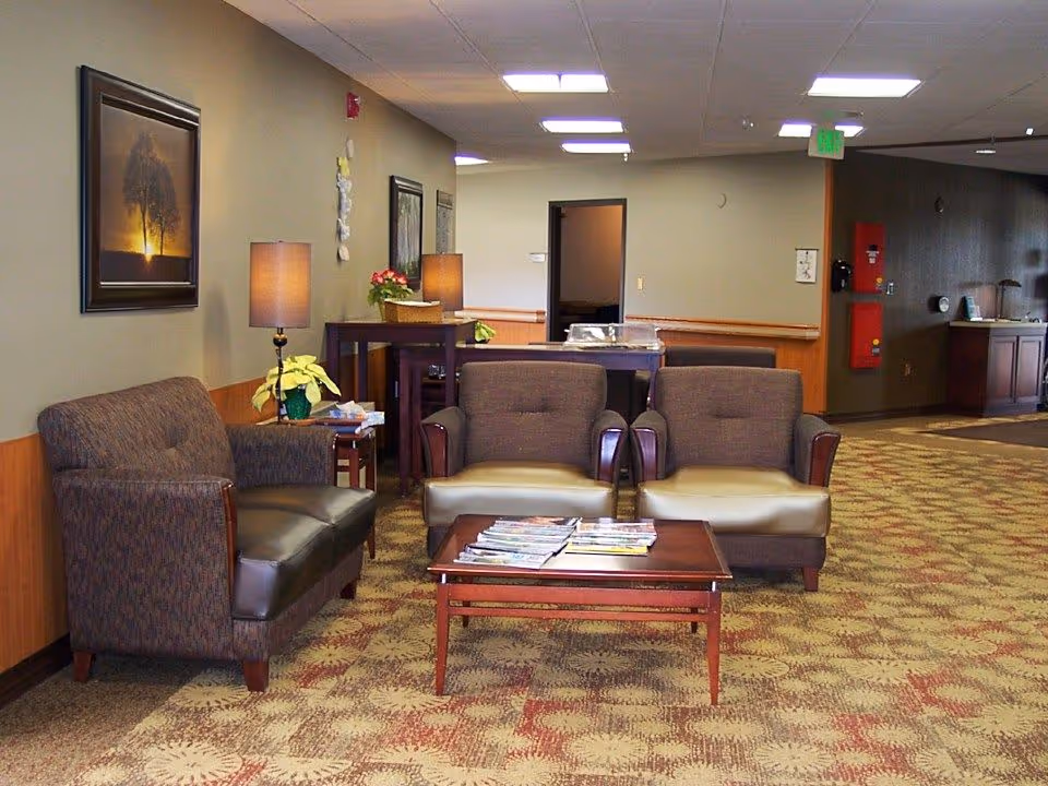 A cozy seating area in a retirement facility with two armchairs and a loveseat arranged around a wooden coffee table holding magazines. The room has patterned carpet, beige walls with framed artwork, two table lamps, and a hallway leading to other rooms.