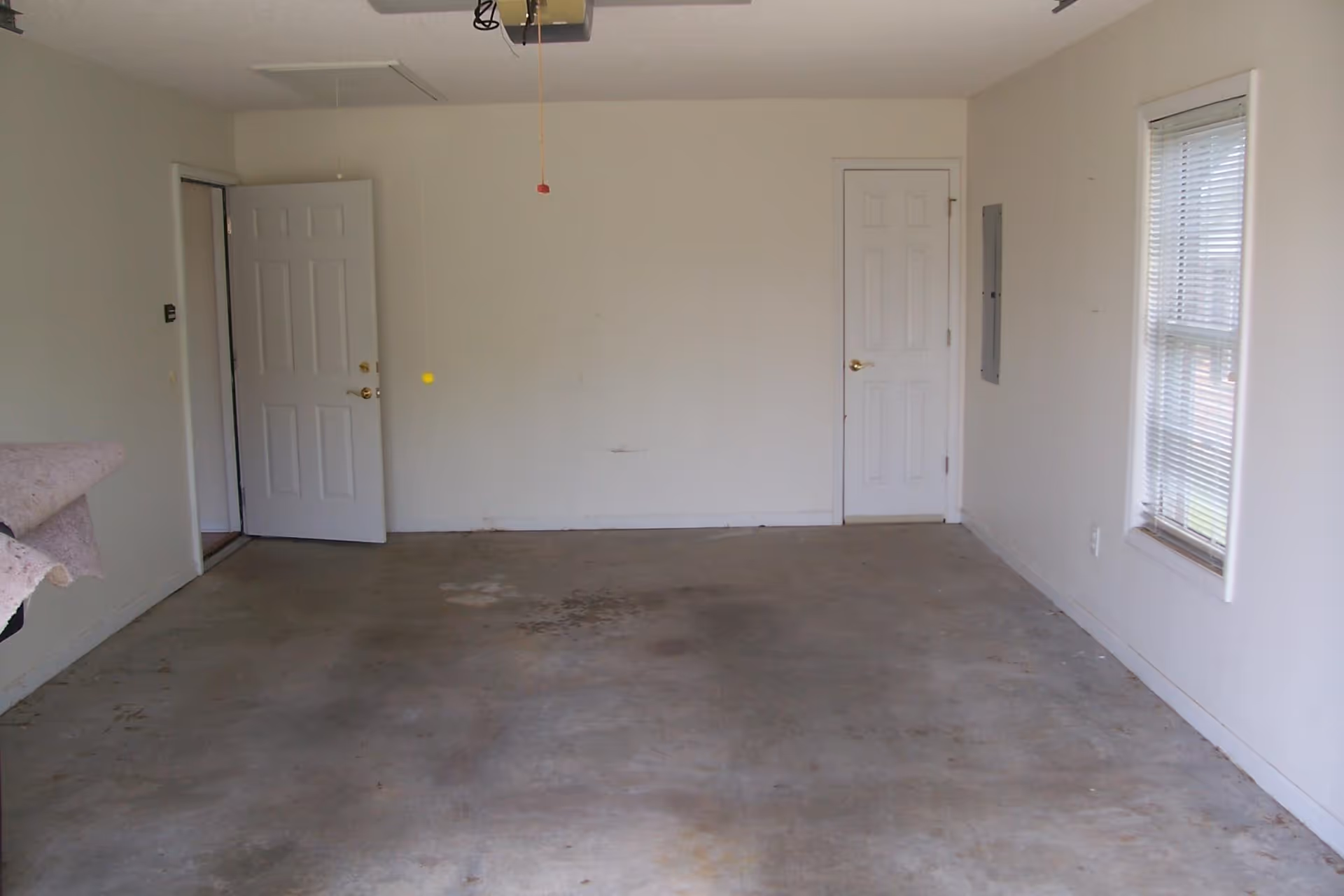 Empty room with concrete floor, white walls, two closed white doors, one open white door, a window with blinds on the right wall, and a ceiling fan pull chain hanging from the ceiling.