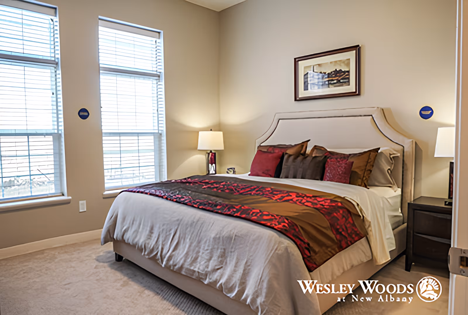 Bright furnished bedroom with a made bed, decorative pillows and runner, bedside lamps, framed art, and two large windows.