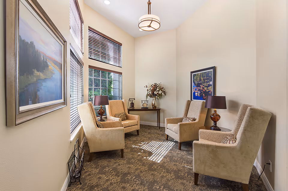 A cozy sitting area with four beige armchairs arranged around two small side tables with lamps. The room has large windows with wooden blinds allowing natural light to enter. There are two framed paintings on the walls and a small table with decorative items and a vase of flowers in the corner. The carpet has a subtle pattern, and the walls are painted a light cream color.