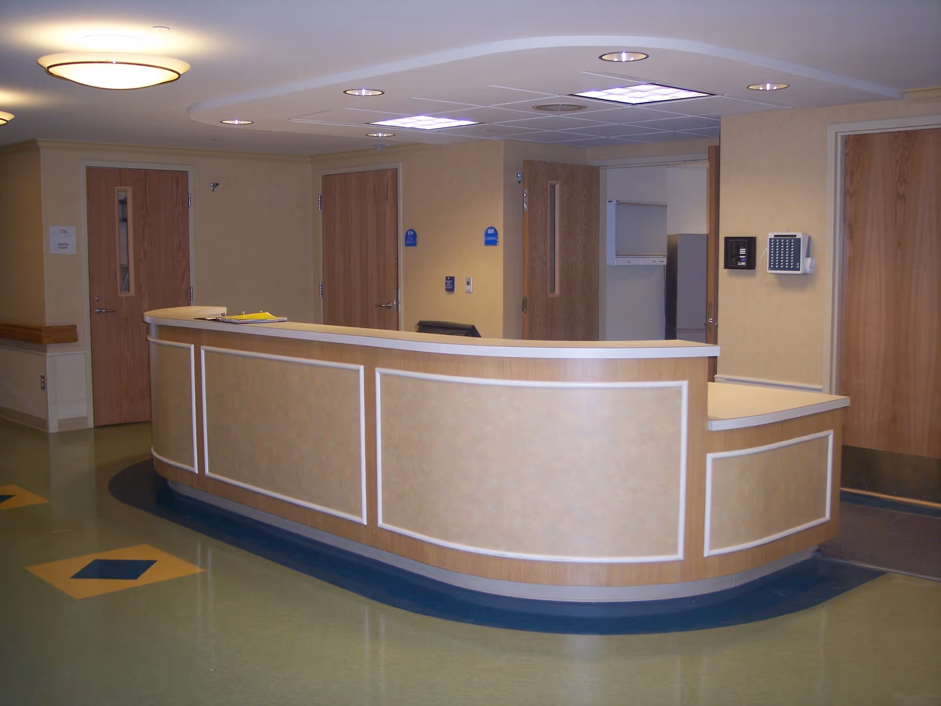 A curved reception desk in a medical or senior care facility with beige paneling and white trim. Behind the desk are several wooden doors, some with small windows. The floor is green with blue and yellow diamond patterns, and the ceiling has recessed lighting and a round ceiling light fixture.