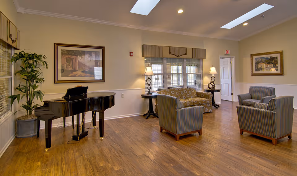 Bright common lounge with a grand piano, sofa and armchairs, side tables with lamps, hardwood floor and skylights.