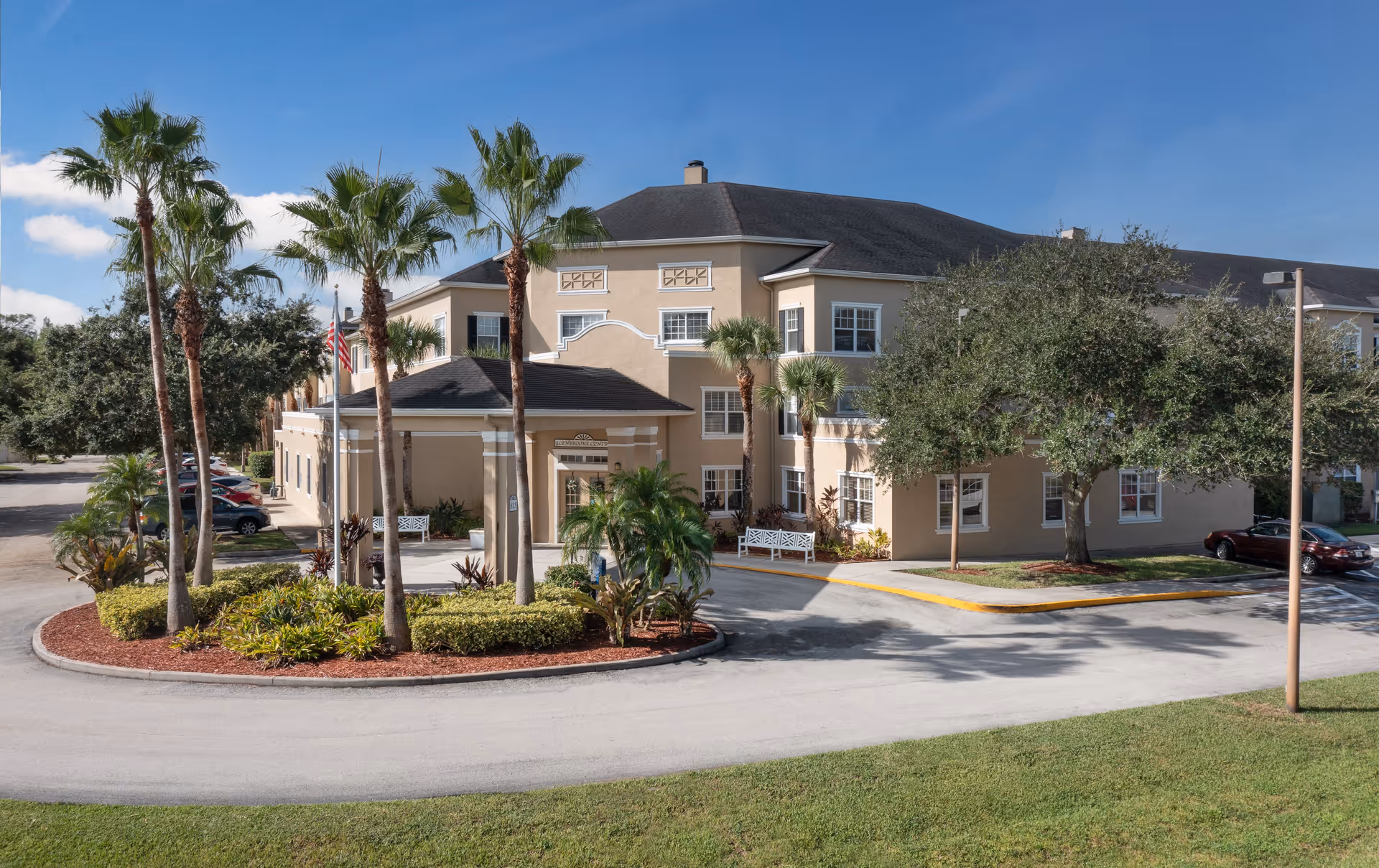 Exterior view of a beige senior living facility building with a dark roof, surrounded by palm trees and other greenery. There is a circular driveway with parked cars and a clear blue sky in the background.