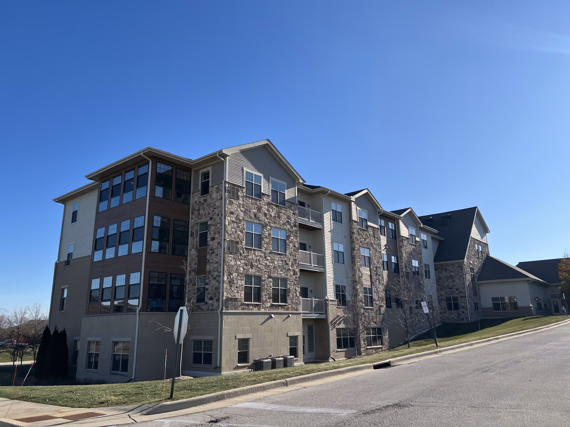 Exterior view of a multi-story senior living facility building with stone and siding facade under a clear blue sky. The building has multiple windows and balconies, with a paved road and grassy area in the foreground.