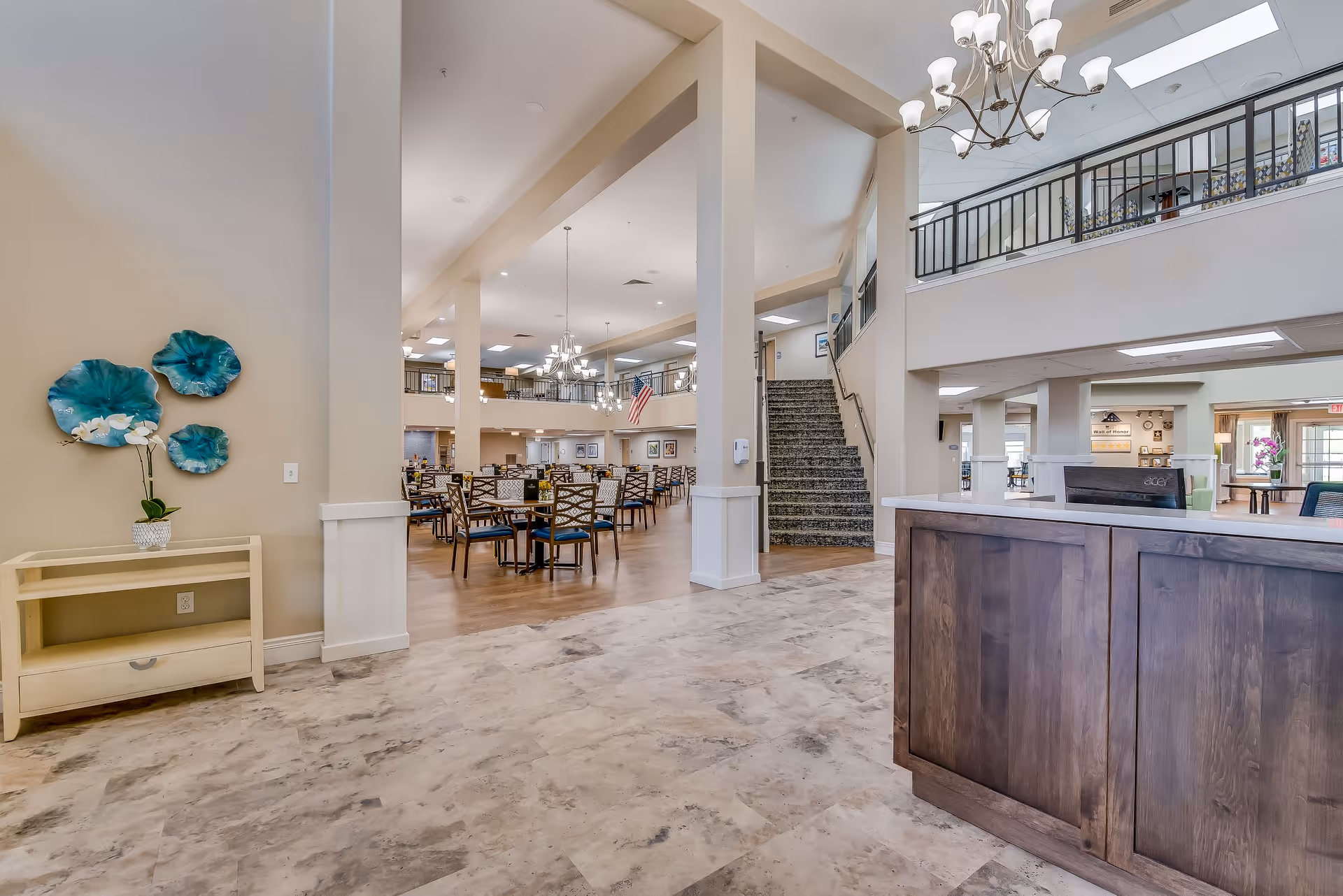 Spacious interior of a senior living facility featuring a reception desk on the right, a staircase leading to an upper level, and a large dining area with multiple tables and chairs in the background. The space is well-lit with chandeliers and natural light, and decorated with blue wall art and a potted orchid on a small table.