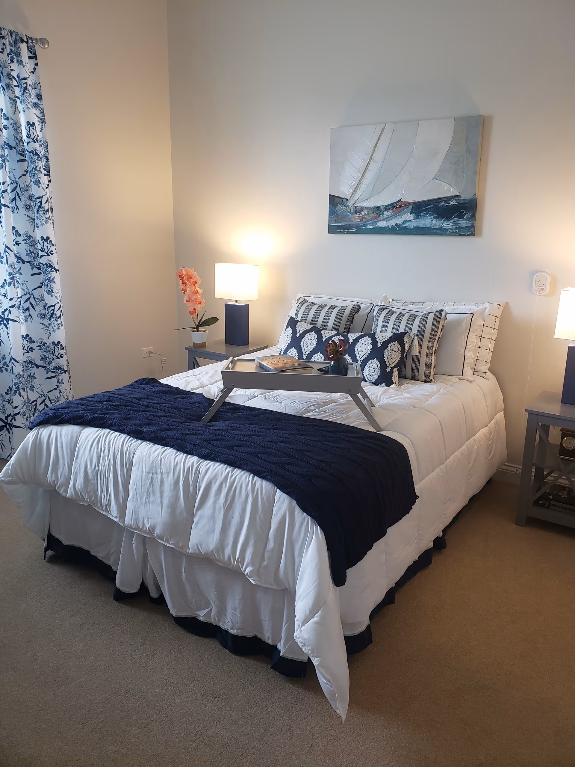 A neatly made bed in a bedroom with white and navy blue bedding, decorative pillows, and a tray on top. There are two bedside tables with lamps, a pink orchid plant on the left table, and a painting of a sailboat on the wall above the bed. Blue and white floral curtains hang on the left side of the room.