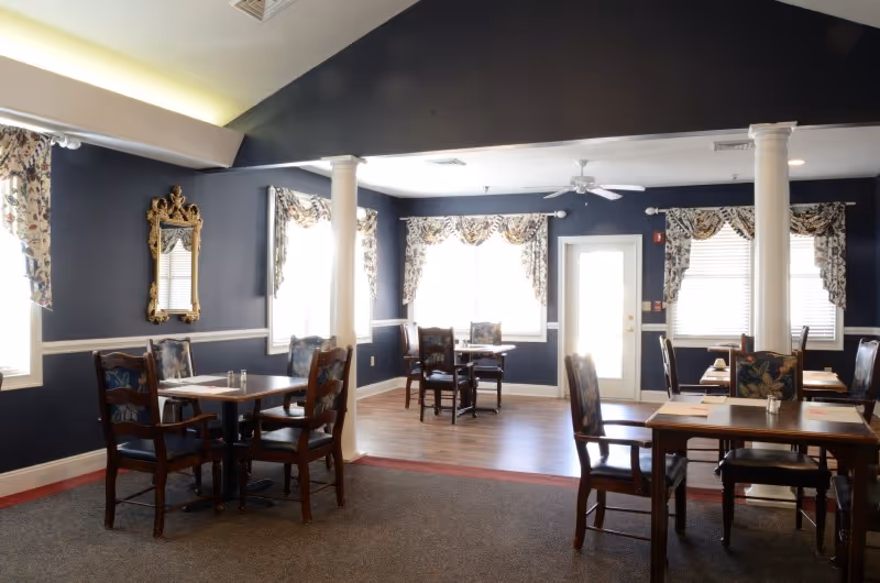 A dining room with several wooden tables and chairs arranged neatly. The walls are painted dark blue with white trim, and there are multiple windows with floral valances letting in natural light. Two white columns stand in the middle of the room, and a decorative mirror hangs on one wall.