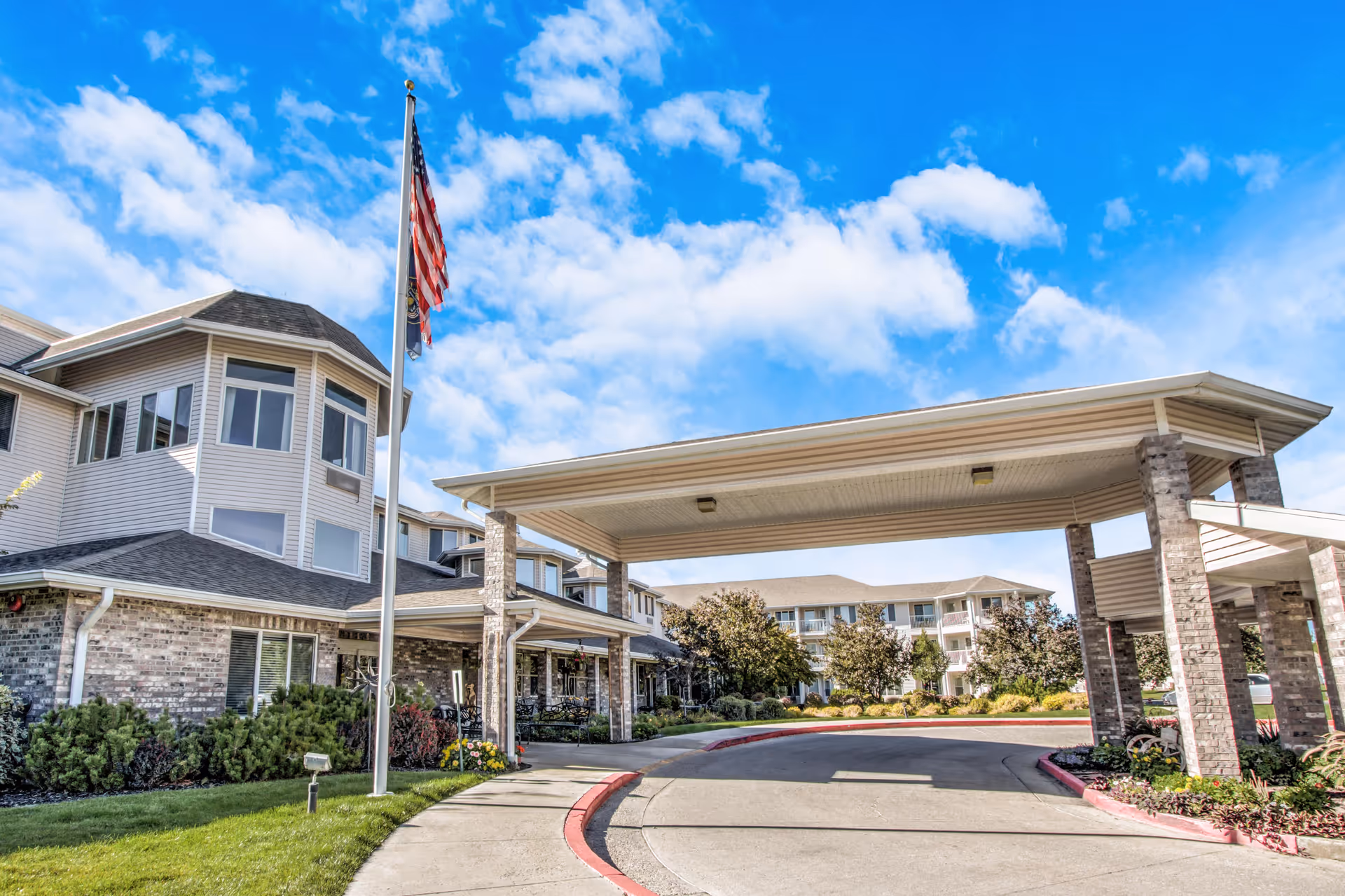 Exterior view of Pioneer Valley Lodge Gracious Retirement Living showing a covered driveway entrance with brick pillars, an American flag on a flagpole, landscaped greenery, and a multi-story building under a partly cloudy blue sky.