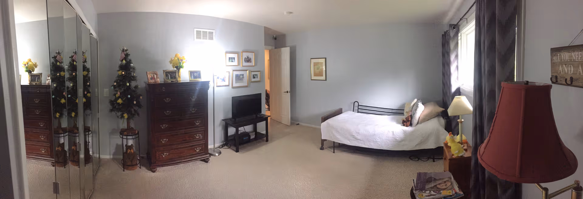A bedroom with light gray walls and beige carpet. The room features a single bed with white bedding and pillows, a wooden dresser with framed photos and a small decorated Christmas tree on top, a TV on a small stand, a floor lamp, and a red lampshade on a side table. There is a mirrored closet door on the left and a window with dark curtains on the right.