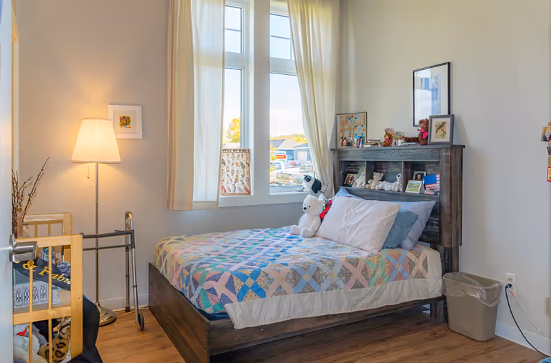 A cozy bedroom in a senior living facility with a bed covered in a colorful quilt, stuffed animals on the bed and headboard, a window with cream curtains letting in natural light, a floor lamp, a walker, and a small trash bin.
