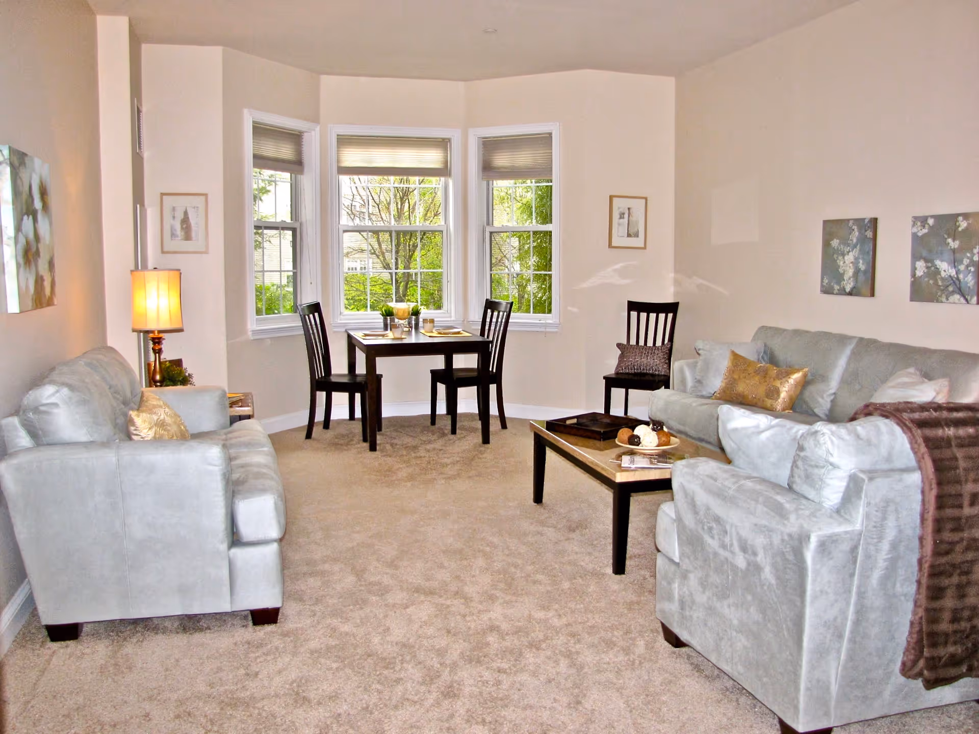 A cozy living room with beige walls and carpet, featuring two light gray sofas and a matching armchair arranged around a wooden coffee table. There is a small dining table with two chairs near a bay window with three large windows showing greenery outside. The room is decorated with framed artwork, a table lamp, and throw pillows.