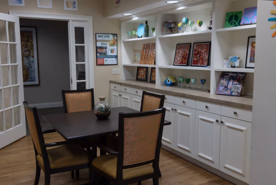 A dining area with a dark wooden table surrounded by four chairs with light brown upholstery. Behind the table is a long white built-in cabinet with multiple drawers and shelves displaying various decorative items including framed pictures, glassware, and small art pieces. The room has light-colored walls and wooden flooring, with French doors on the left side leading to another room.