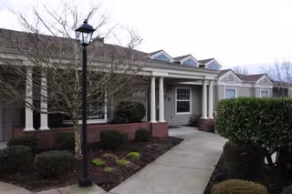 Exterior view of a single-story senior living facility building with a covered entrance supported by white columns, a brick planter, a sidewalk leading to the entrance, a street lamp, and leafless trees and bushes around the building.