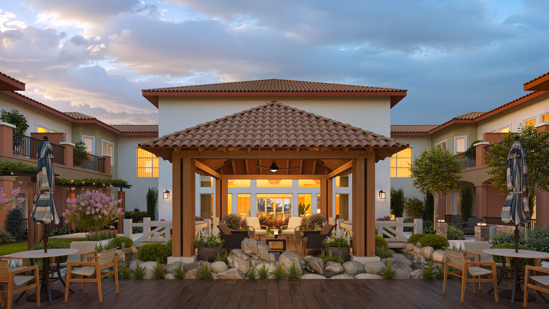 Outdoor courtyard area of a senior living facility with a central wooden gazebo surrounded by rocks and plants. The gazebo has seating with chairs and a table inside. Around the courtyard are two-story buildings with balconies, windows lit warmly from inside, and patio tables with umbrellas. The sky is partly cloudy during sunset.