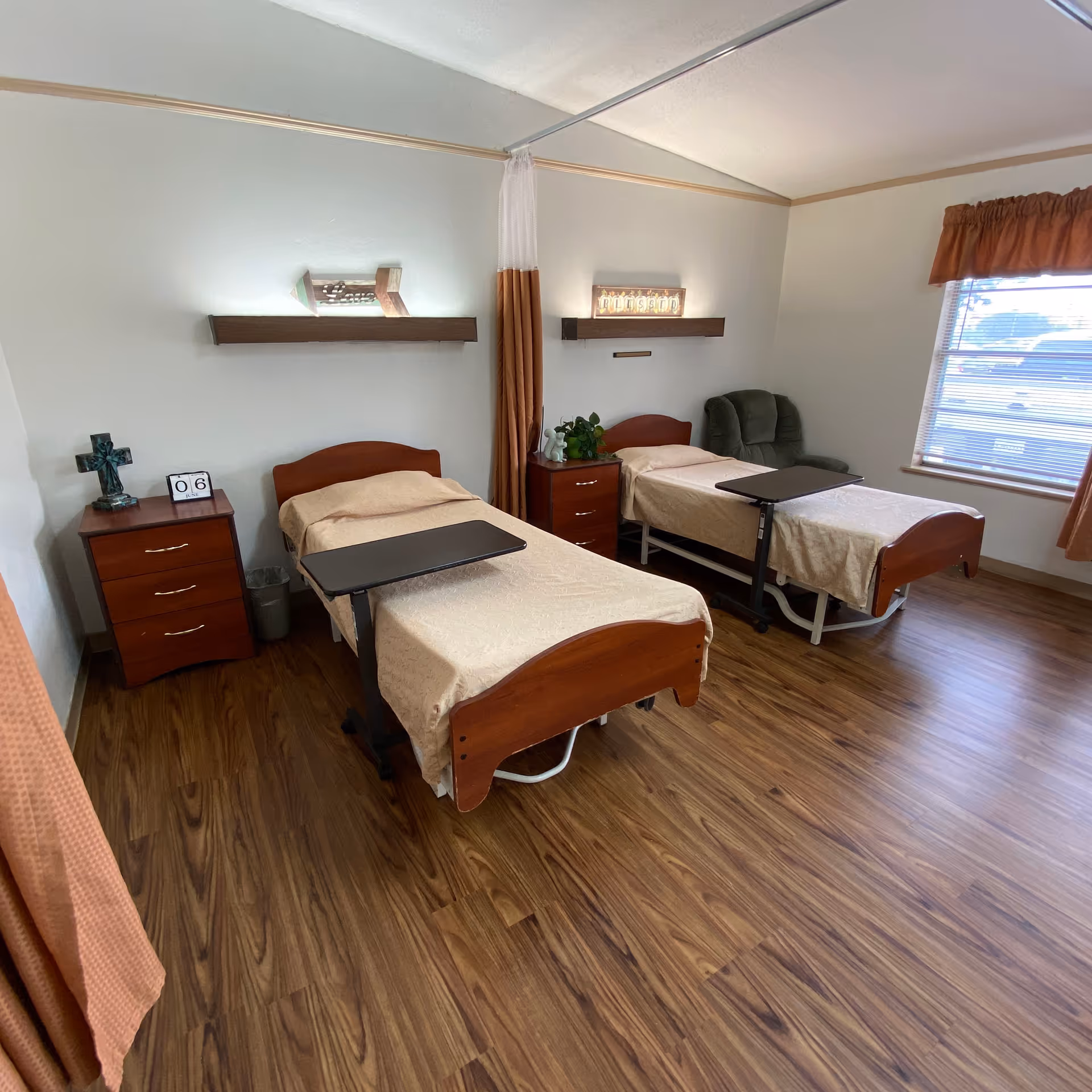 A nursing facility bedroom with two single beds, each with a beige bedspread and an overbed table. There are two wooden nightstands between and beside the beds, one with a decorative cross and a calendar block showing the number 06. A green armchair is positioned near the window with orange curtains, and the room has wood flooring and white walls.