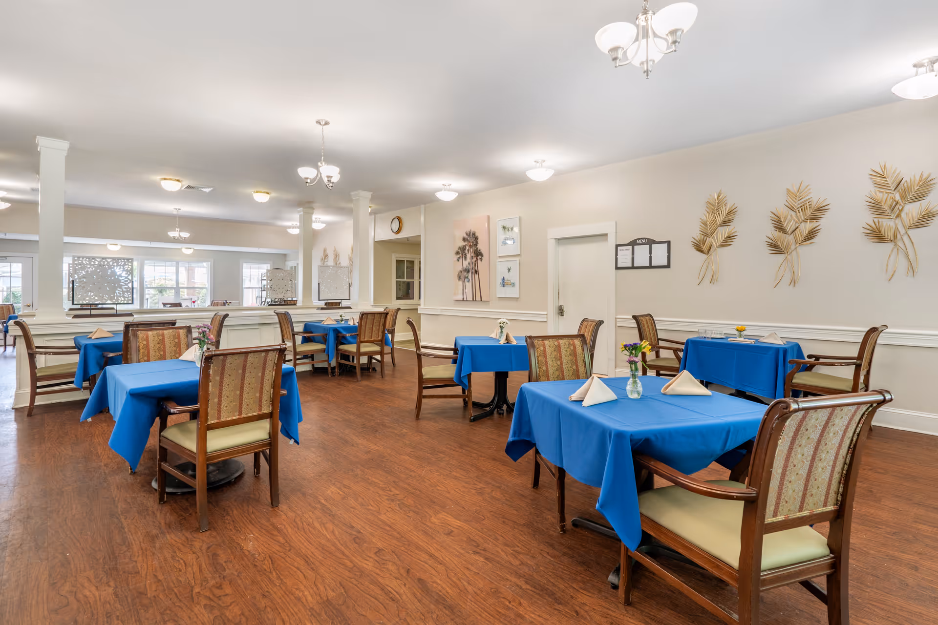A dining room with several tables covered in blue tablecloths, each set with folded napkins and small flower vases. The room has wooden flooring, beige walls decorated with framed pictures and gold leaf wall art, and multiple ceiling light fixtures providing bright illumination.