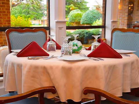 A round dining table set for four with a white tablecloth, red folded napkins, upside-down wine glasses, plates, and silverware. The table is in a room with large windows showing green bushes and trees outside, and there are upholstered chairs with wooden arms around the table.