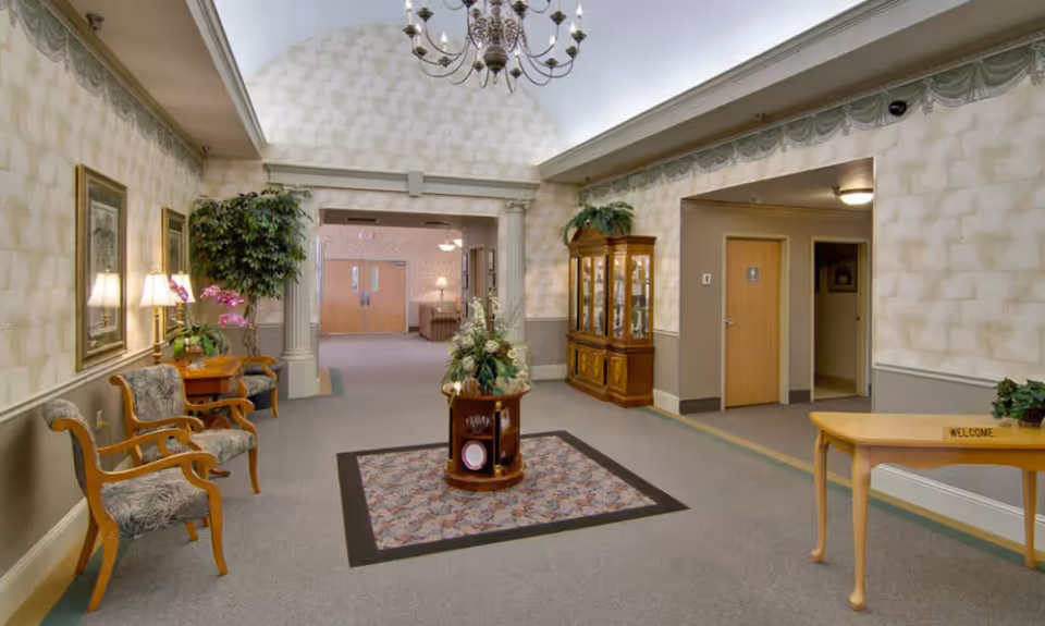 Spacious decorated lobby of a senior living facility with seating, a central floral display, chandelier, and a welcome table.