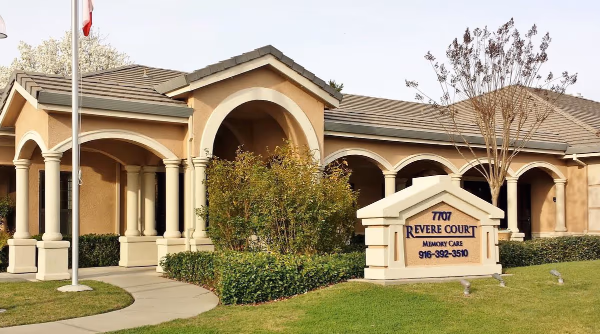 Exterior view of Revere Court Memory Care facility showing a beige building with arched columns, a flagpole, landscaped bushes, and a sign with the facility's name and phone number on a grassy lawn.