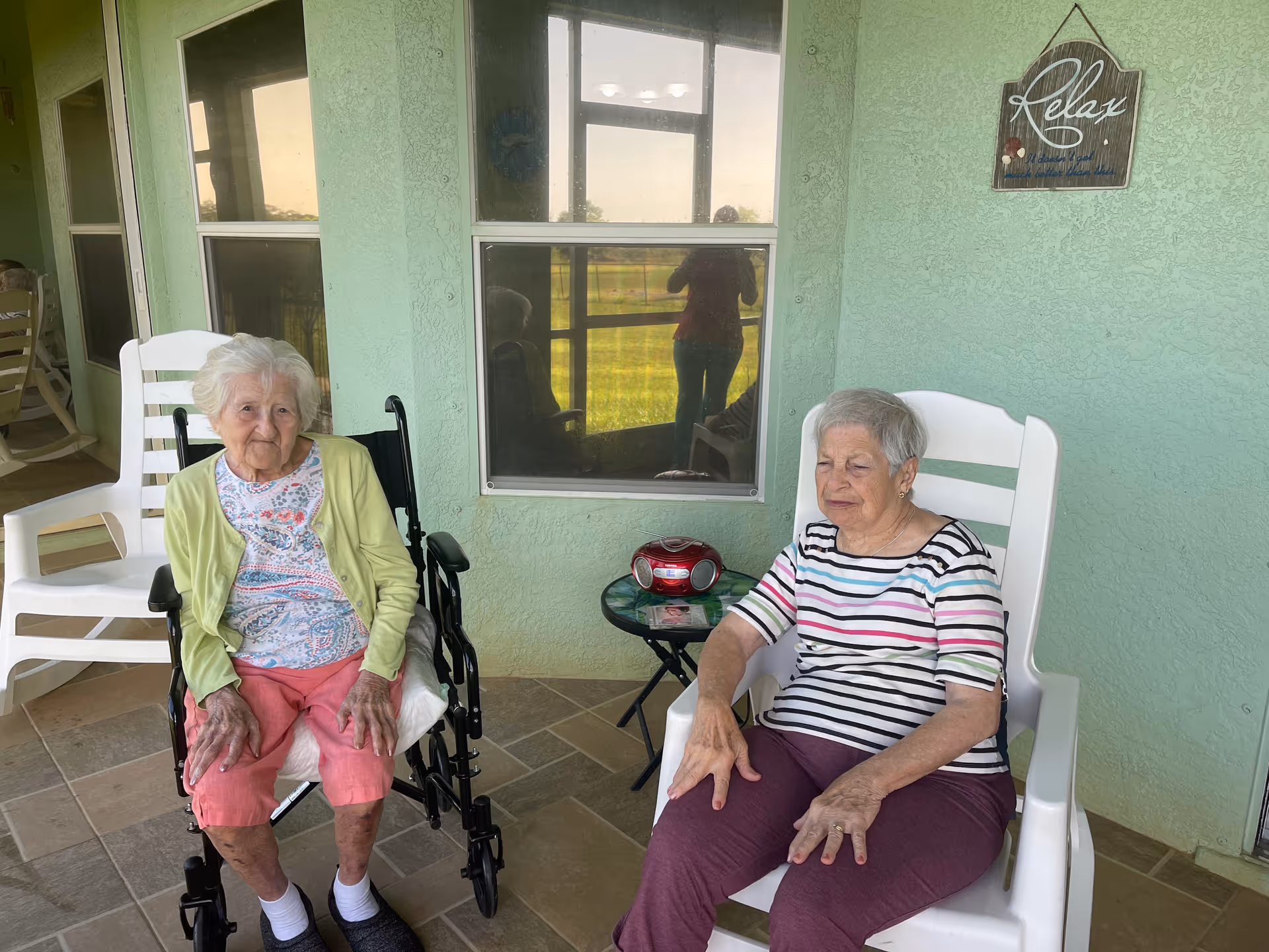 Two elderly women sitting outside on a tiled patio. One woman is seated in a wheelchair wearing a light green cardigan and pink pants, while the other woman is sitting in a white plastic chair wearing a striped shirt and purple pants. Behind them is a green wall with a window and a small table holding a red radio. A decorative sign on the wall reads 'Relax'.