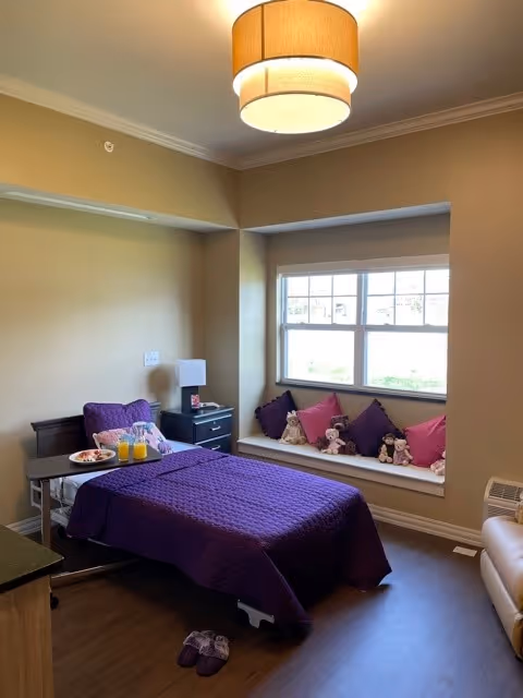 A cozy bedroom with a single bed covered in a purple quilt, a window seat lined with pillows and stuffed animals, a nightstand and overhead light.