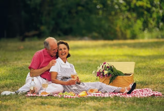 An elderly couple enjoying a picnic on a red and white checkered blanket in a grassy outdoor area. The man in a pink shirt is sitting behind the woman in a white shirt and checkered overalls, both smiling and holding drinks. There is a picnic basket with flowers and food items around them.