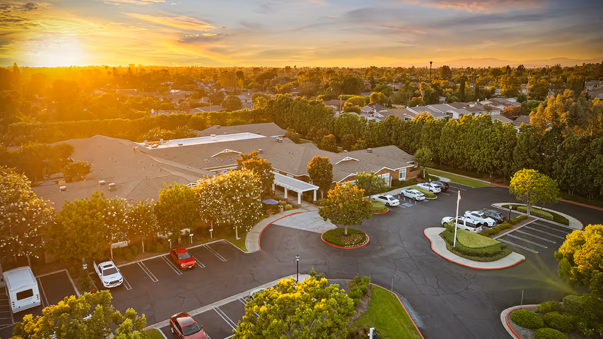 Aerial view of Ivy Park at Tustin senior living facility at sunset, showing the building surrounded by trees, parked cars, and a well-maintained parking lot with landscaped islands.