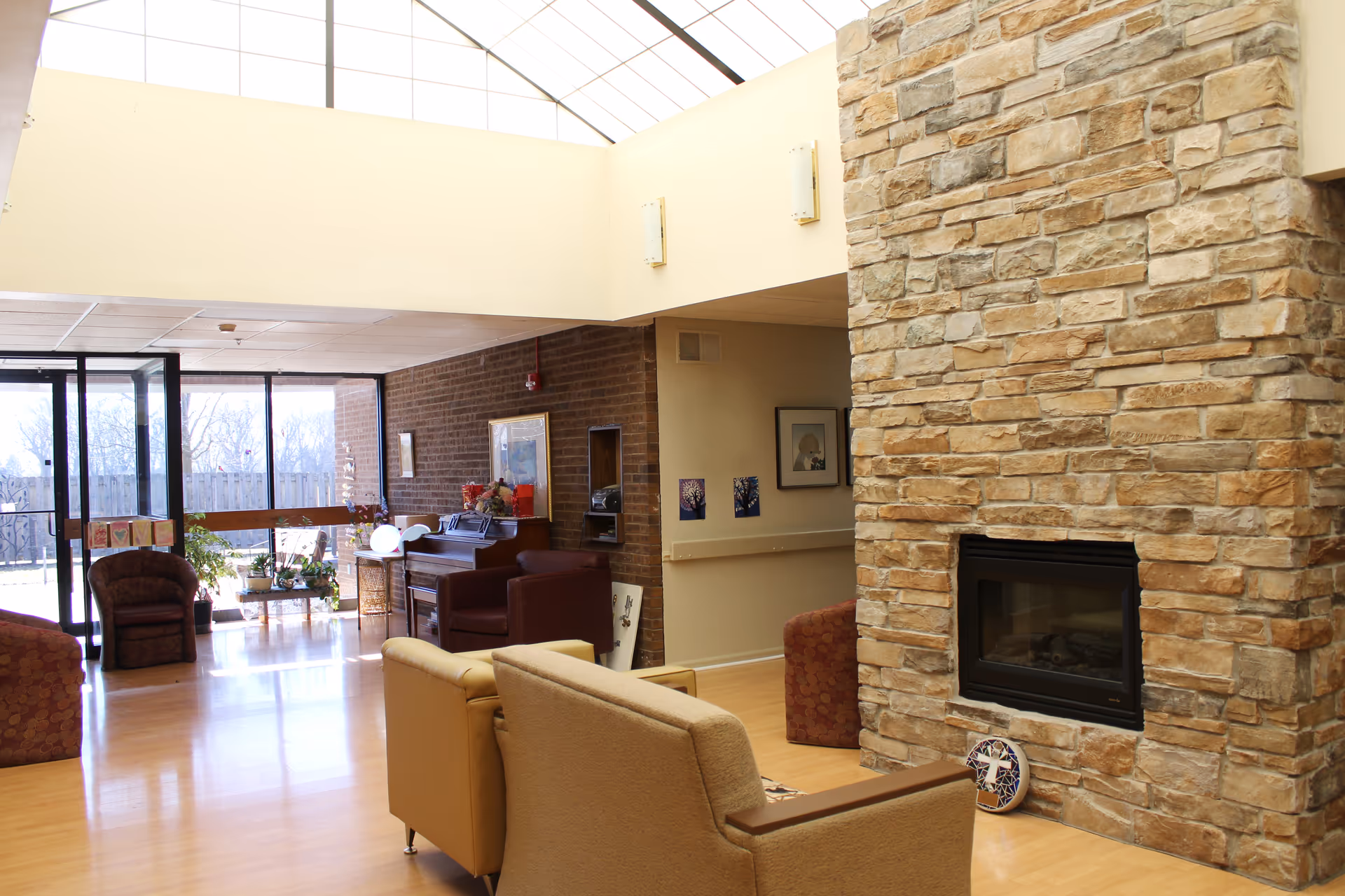 A bright and spacious living room area with a large stone fireplace on the right. The room features comfortable seating including beige and patterned armchairs, a piano against a brick wall, and large windows letting in natural light. There are plants and decorative items near the windows, and artwork on the walls.