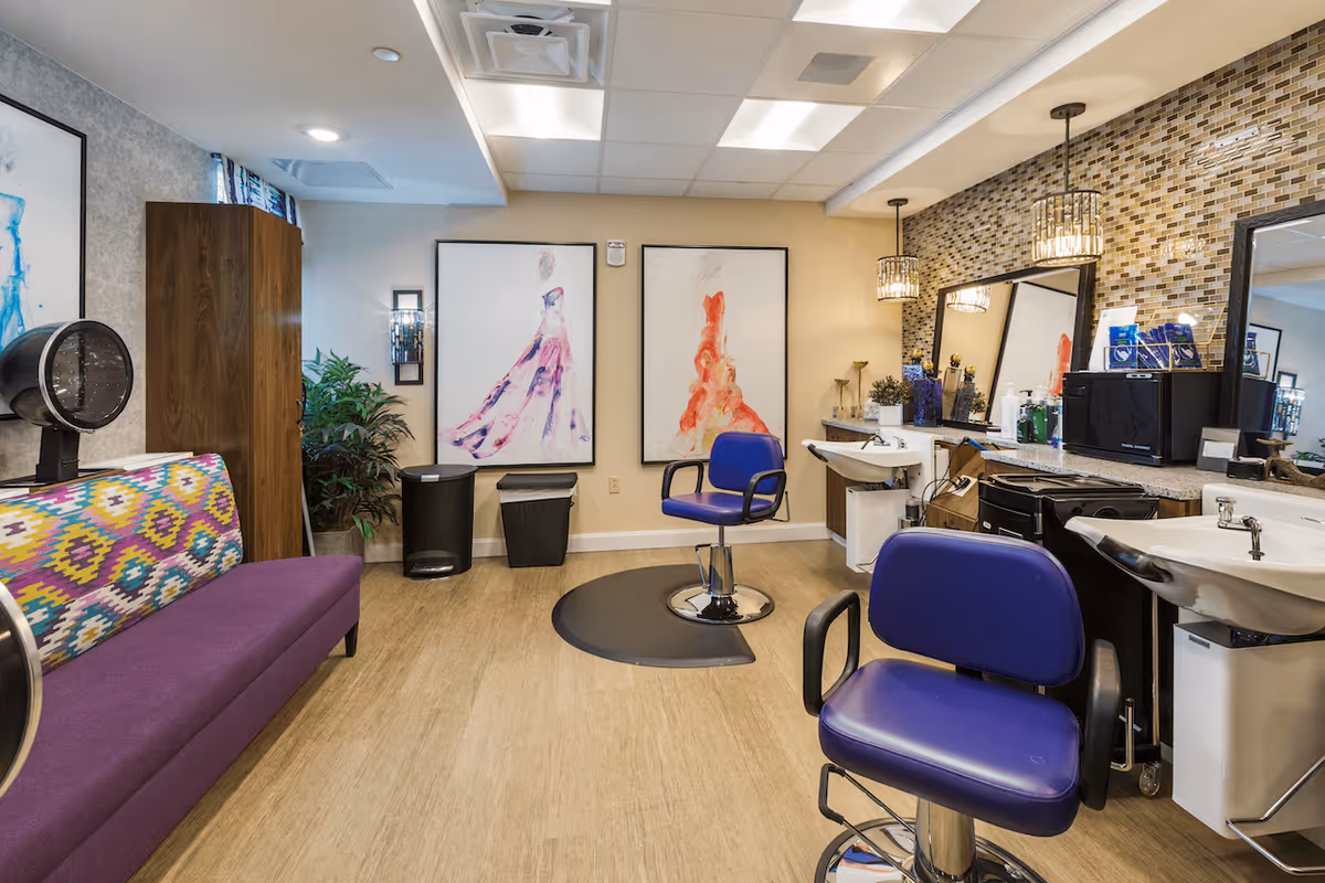 Interior of a salon area in a senior living facility with two purple salon chairs, hair washing sinks, a colorful patterned bench, large framed artwork on the wall, and a wooden cabinet. The room has a tiled backsplash behind the sinks and mirrors, with various hair care products on the counter.